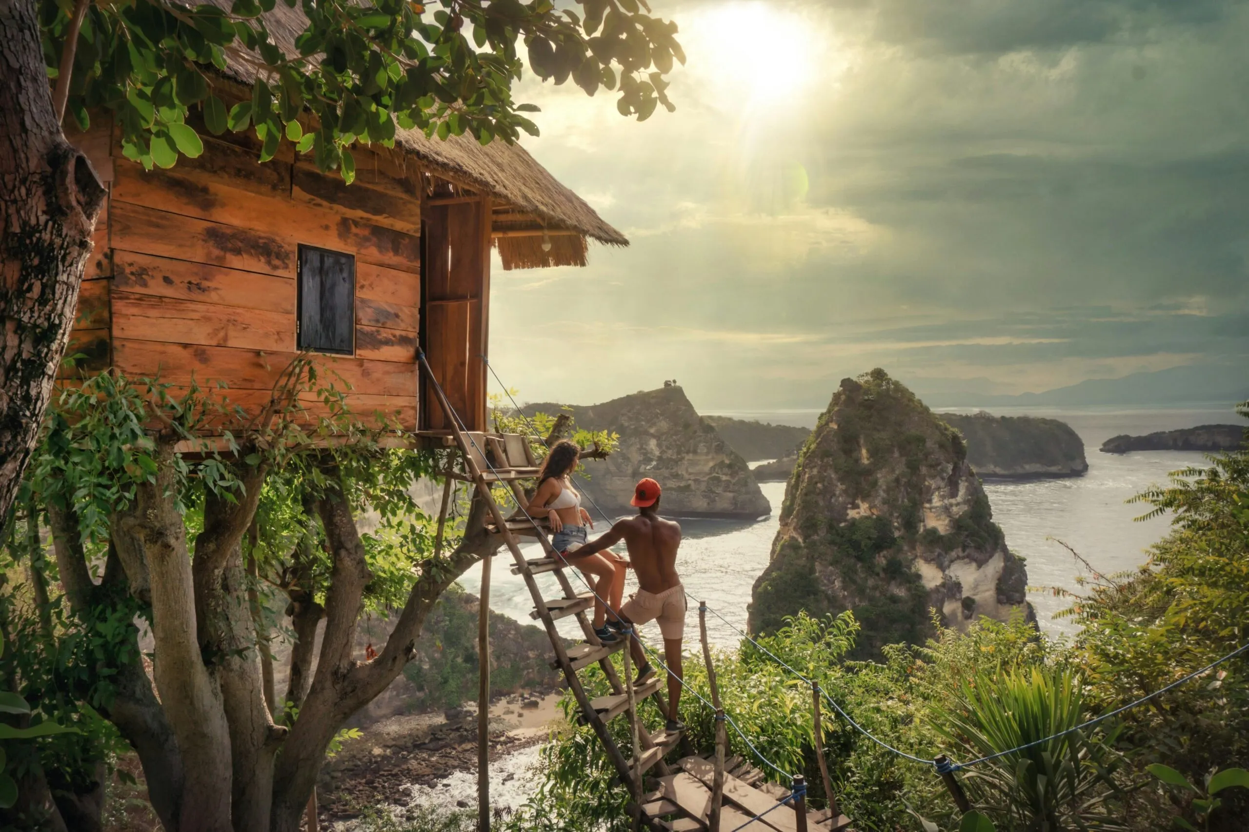 Two travelers climbing wooden steps to a treehouse above a rocky coastline