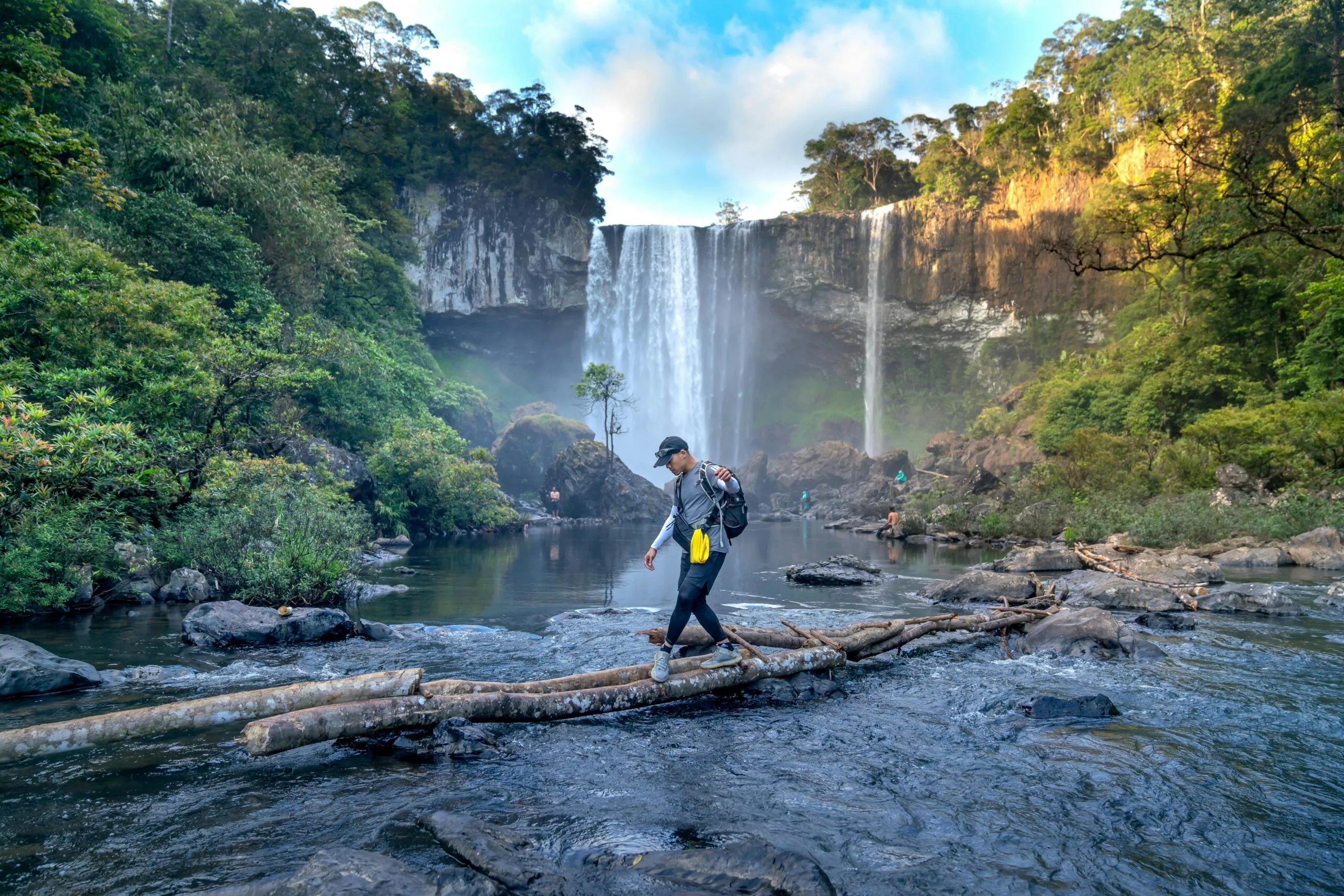 Hiker crossing a log in a river below a jungle waterfall